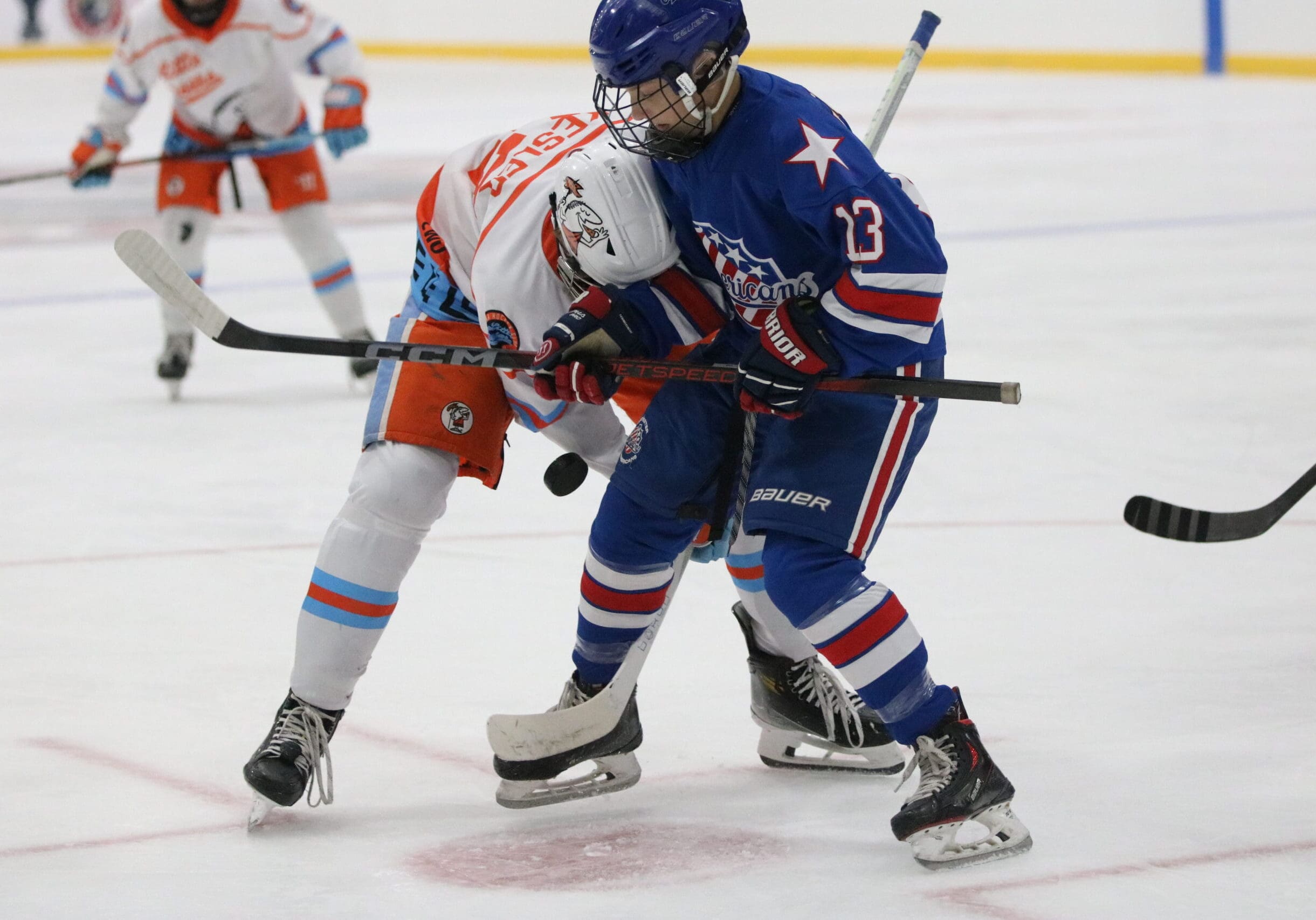 Rochester Jr. Americans player in blue jersey #13 battles for puck during faceoff against Little Caesars Hockey Club player in white and orange jersey during AAA youth hockey game.